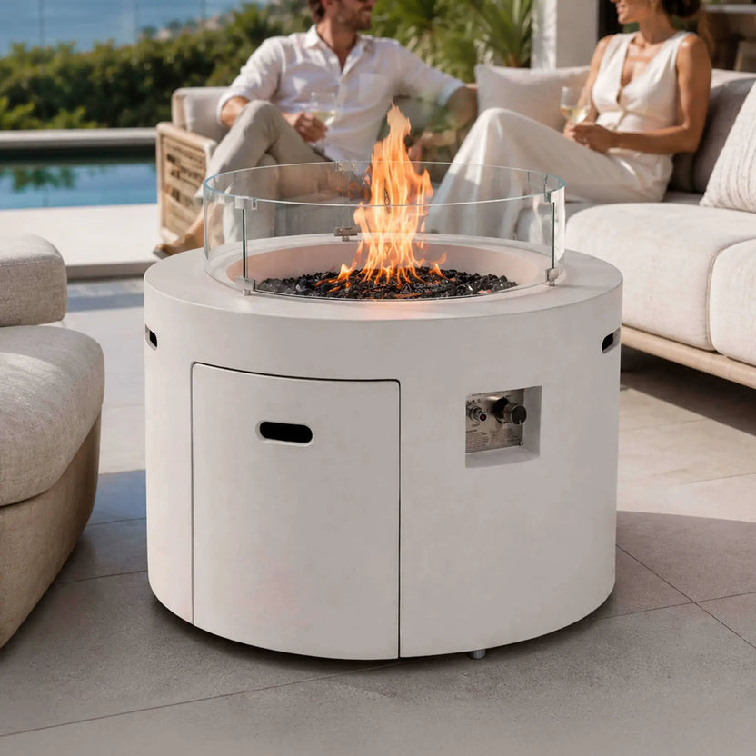 Lifestyle image of the Flame Havens Refuge Series 36-inch Round Propane Gas GFRC Concrete Fire Pit in Ivory White featuring premium Black Reflective Fire Glass and an optional tempered glass wind guard. Positioned on a luxury outdoor patio beside comfortable seating, the fire pit creates a warm focal point while complementing elegant modern outdoor living spaces.