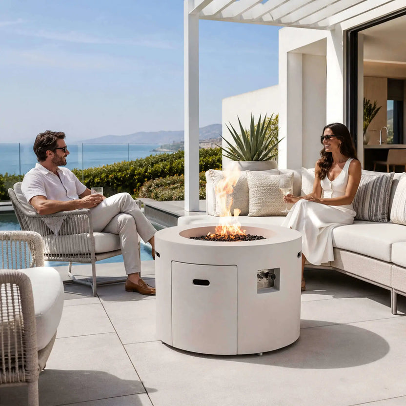 Lifestyle image of the Flame Havens Refuge Series 36-inch Round Propane Gas GFRC Concrete Fire Pit in Ivory White with Black Reflective Fire Glass and a beautiful natural flame creating an inviting centerpiece on a luxury outdoor patio. A couple relaxes nearby with drinks overlooking the pool, showcasing how this propane gas fire pit enhances outdoor entertaining, comfort, and modern backyard living.