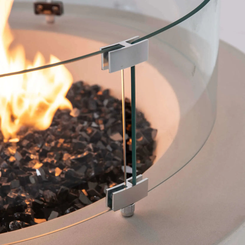 Close-up detail of the optional tempered glass wind guard mounted on the Flame Havens Refuge Series 36-inch Round Propane Gas GFRC Concrete Fire Pit in Ivory White. The image highlights the precision stainless steel mounting hardware, polished glass edge, premium Black Reflective Fire Glass, and natural propane flame, showcasing the quality craftsmanship and secure wind guard installation.