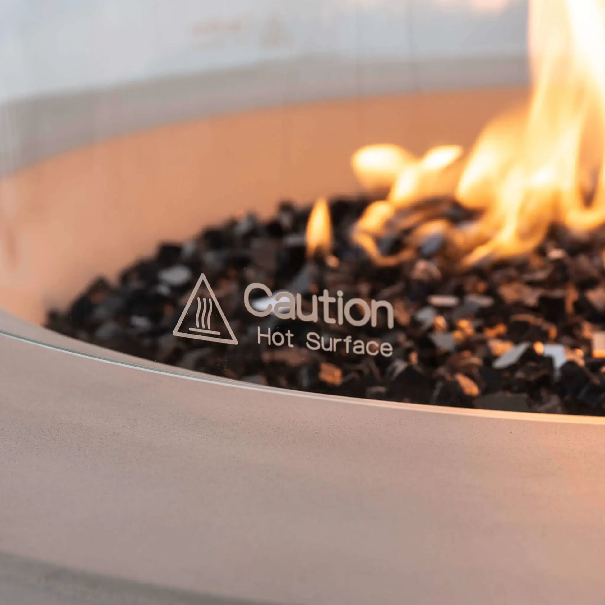 Close-up of the tempered glass wind guard on the Flame Havens Refuge Series 36-inch Round Propane Gas GFRC Concrete Fire Pit in Ivory White, featuring the etched "Caution Hot Surface" safety marking. The image also showcases the vibrant propane flame, premium Black Reflective Fire Glass, and smooth GFRC concrete rim, emphasizing both safety and refined outdoor fire pit design.