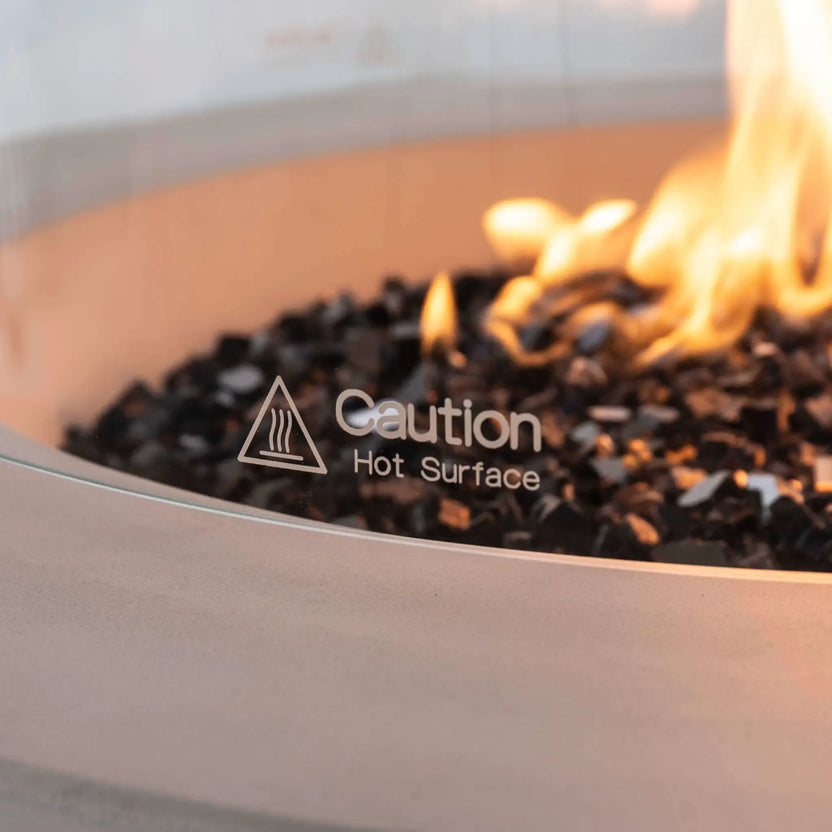 Close-up of the tempered glass wind guard on the Flame Havens Refuge Series 36-inch Round Propane Gas GFRC Concrete Fire Pit in Ivory White, featuring the etched "Caution Hot Surface" safety marking. The image also showcases the vibrant propane flame, premium Black Reflective Fire Glass, and smooth GFRC concrete rim, emphasizing both safety and refined outdoor fire pit design.