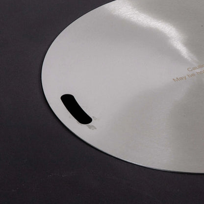Detailed close-up of the Flame Havens Refuge Series 43-inch Round Natural Gas Black GFRC Concrete Fire Pit stainless steel burner cover. The brushed stainless steel lid sits flush within the fire pit opening and features integrated lifting slots, providing a sleek tabletop appearance while protecting the burner when not in use.