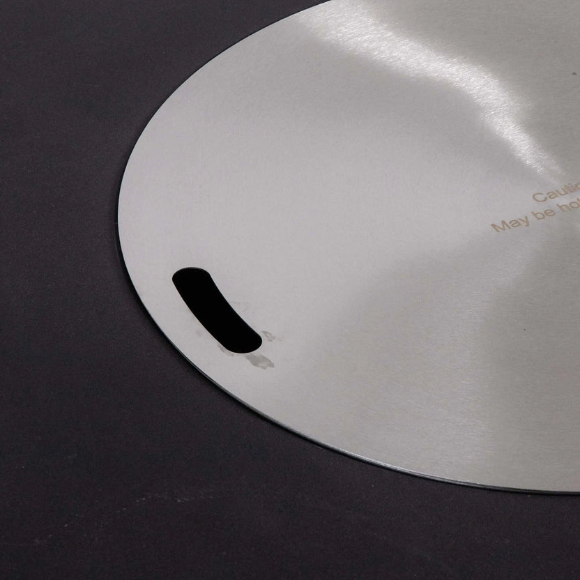 Detailed close-up of the Flame Havens Refuge Series 43-inch Round Natural Gas Black GFRC Concrete Fire Pit stainless steel burner cover. The brushed stainless steel lid sits flush within the fire pit opening and features integrated lifting slots, providing a sleek tabletop appearance while protecting the burner when not in use.