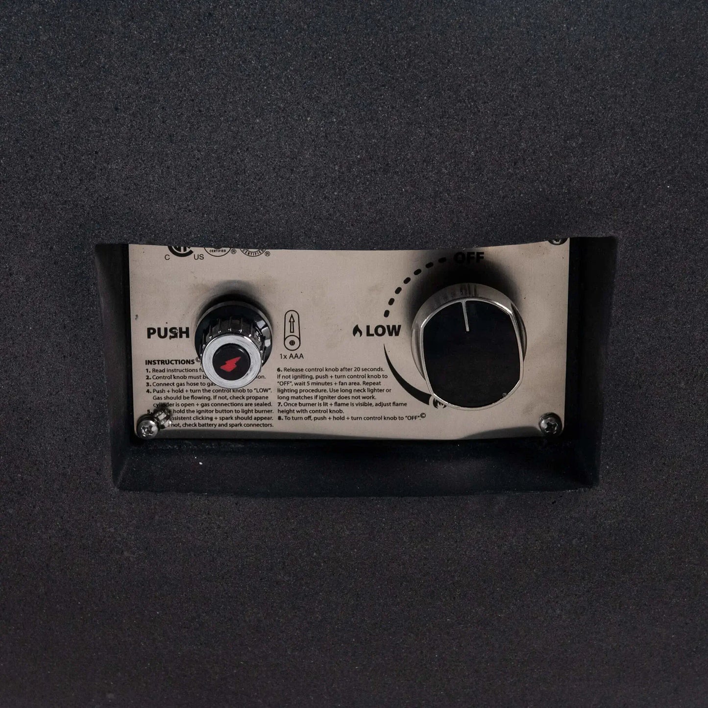 Close-up of the control panel on the Flame Havens Refuge Series 43-inch Round Natural Gas Black GFRC Concrete Fire Pit. The recessed stainless steel panel features the CSA-certified push-button ignition, flame control knob, and operating instructions, providing convenient and reliable ignition while preserving the fire pit's streamlined appearance.