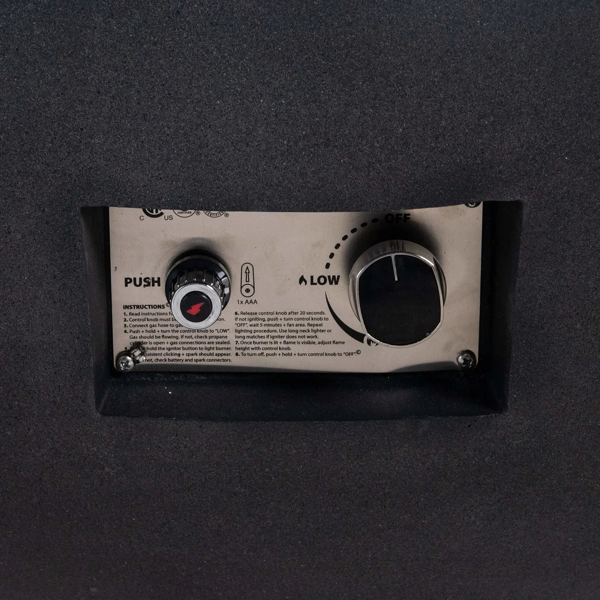 Close-up of the control panel on the Flame Havens Refuge Series 43-inch Round Natural Gas Black GFRC Concrete Fire Pit. The recessed stainless steel panel features the CSA-certified push-button ignition, flame control knob, and operating instructions, providing convenient and reliable ignition while preserving the fire pit's streamlined appearance.