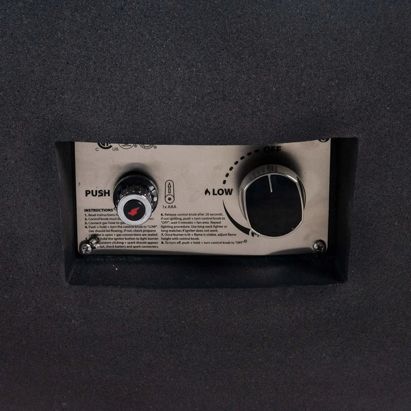 Close-up of the control panel on the Flame Havens Refuge Series 43-inch Round Natural Gas Black GFRC Concrete Fire Pit. The recessed stainless steel panel features the CSA-certified push-button ignition, flame control knob, and operating instructions, providing convenient and reliable ignition while preserving the fire pit's streamlined appearance.