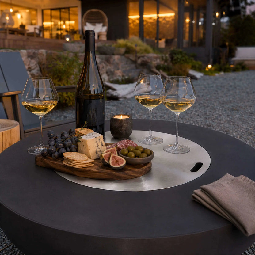 The Flame Havens Refuge Series 43-inch Round Natural Gas Black GFRC Concrete Fire Pit with its stainless steel burner cover installed and styled as an elegant outdoor coffee table. A charcuterie board, wine glasses, candle, and bottle create an inviting entertaining space, showcasing the fire pit's dual-purpose functionality for luxury outdoor living.