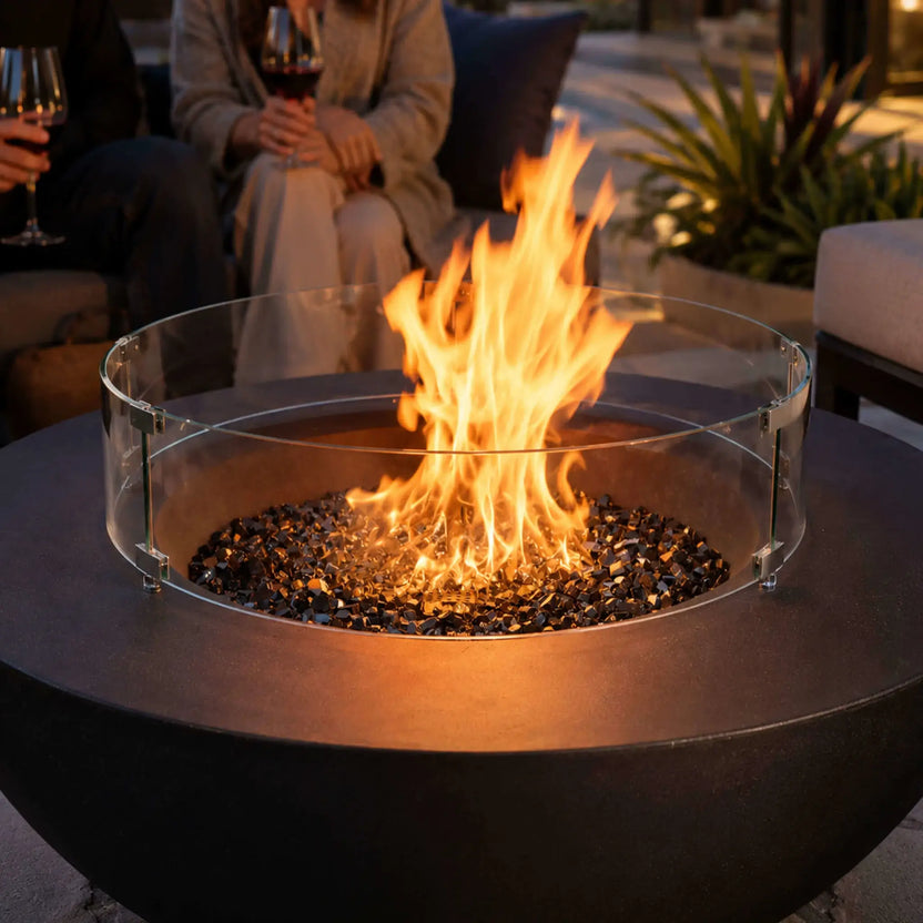 Lifestyle image of the Flame Havens Refuge Series 43-inch Round Natural Gas Black GFRC Concrete Fire Pit featuring reflective tempered black fire glass and a tempered glass wind guard. Set within a luxurious outdoor seating area at dusk, the glowing fire creates a warm, inviting atmosphere for entertaining and relaxing.