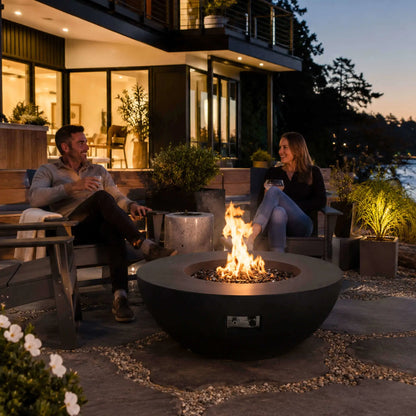 Lifestyle view of the Flame Havens Refuge Series 43-inch Round Natural Gas Black GFRC Concrete Fire Pit in use outside a contemporary home. A couple relaxes around the fire while enjoying drinks, highlighting the reflective tempered black fire glass, warm flame, and sophisticated design that enhances luxury outdoor living and social gatherings.