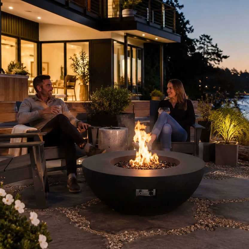 Lifestyle view of the Flame Havens Refuge Series 43-inch Round Natural Gas Black GFRC Concrete Fire Pit in use outside a contemporary home. A couple relaxes around the fire while enjoying drinks, highlighting the reflective tempered black fire glass, warm flame, and sophisticated design that enhances luxury outdoor living and social gatherings.