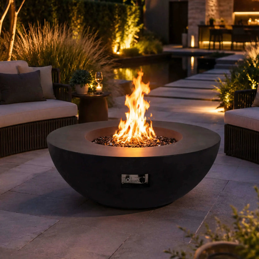 The Flame Havens Refuge Series 43-inch Round Natural Gas Black GFRC Concrete Fire Pit displayed in a luxury backyard patio with reflective tempered black fire glass and a lively flame. Positioned between comfortable outdoor seating beside a modern pool, the fire pit creates a welcoming centerpiece for evening entertaining.