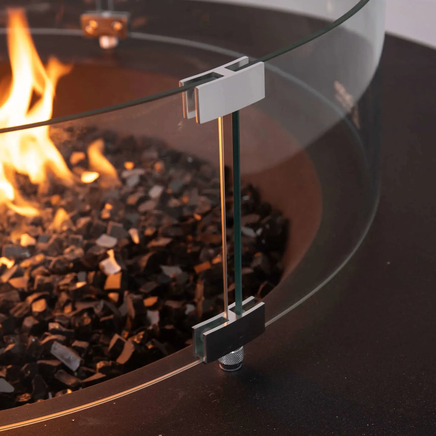 Close-up of the tempered glass wind guard on the Flame Havens Refuge Series 43-inch Round Natural Gas Fire Pit. The image highlights the polished stainless steel mounting brackets, adjustable support hardware, and thick tempered glass surrounding the reflective black fire glass and flame for enhanced safety and premium craftsmanship.