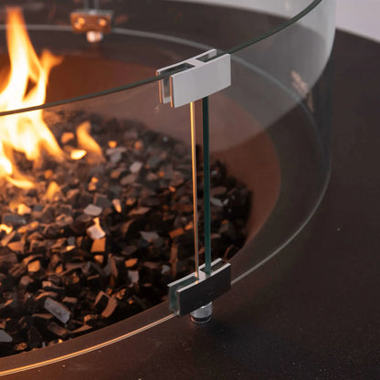 Close-up of the tempered glass wind guard on the Flame Havens Refuge Series 43-inch Round Natural Gas Fire Pit. The image highlights the polished stainless steel mounting brackets, adjustable support hardware, and thick tempered glass surrounding the reflective black fire glass and flame for enhanced safety and premium craftsmanship.