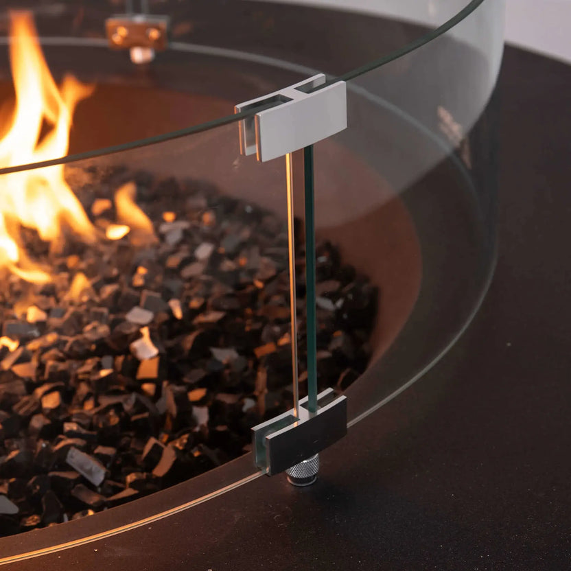 Close-up of the tempered glass wind guard on the Flame Havens Refuge Series 43-inch Round Natural Gas Fire Pit. The image highlights the polished stainless steel mounting brackets, adjustable support hardware, and thick tempered glass surrounding the reflective black fire glass and flame for enhanced safety and premium craftsmanship.