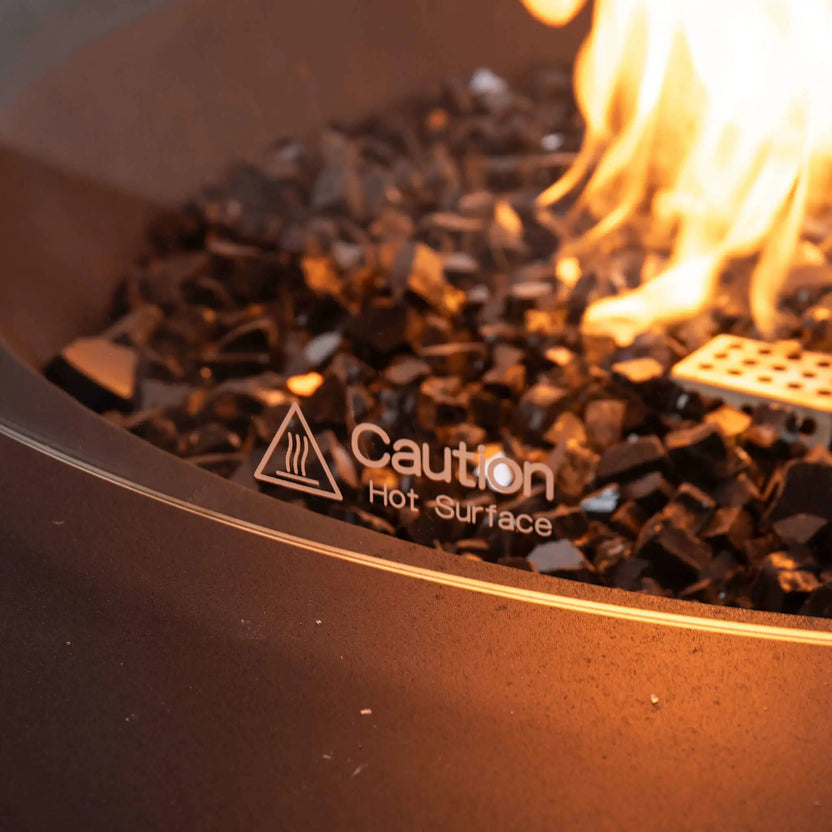Detailed close-up of the Flame Havens Refuge Series tempered glass wind guard showing the etched "Caution Hot Surface" safety marking. Reflective tempered black fire glass and an active natural gas flame are visible behind the clear glass, showcasing the premium safety features and elegant finish of the 43-inch round fire pit.