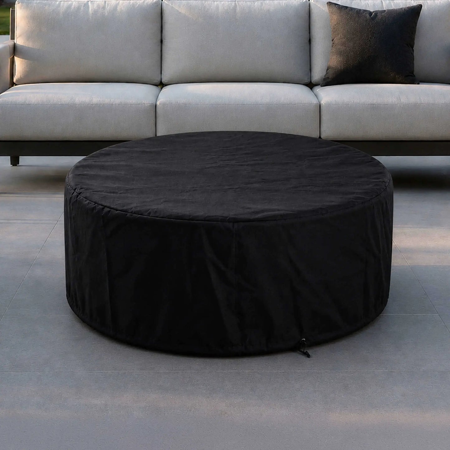Lifestyle image of the Flame Havens Refuge Series 43-inch Round Natural Gas Gray GFRC Concrete Fire Pit covered with the fitted black canvas fabric cover in front of a modern outdoor sofa. The protective cover blends seamlessly into a luxury patio while helping preserve the fire pit during periods of non-use.