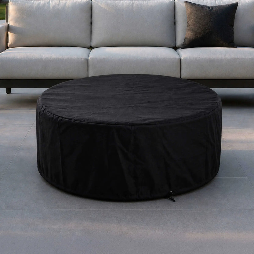 Lifestyle image of the Flame Havens Refuge Series 43-inch Round Natural Gas Gray GFRC Concrete Fire Pit covered with the fitted black canvas fabric cover in front of a modern outdoor sofa. The protective cover blends seamlessly into a luxury patio while helping preserve the fire pit during periods of non-use.