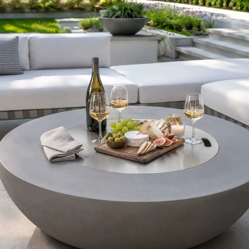 Lifestyle image of the Flame Havens Refuge Series 43-inch Round Natural Gas Gray GFRC Concrete Fire Pit with the stainless steel burner cover in place, serving as an outdoor coffee table. The surface is styled with wine, cheese, fruit, crackers, and candles, demonstrating how the fire pit becomes a functional centerpiece when the burner is covered.