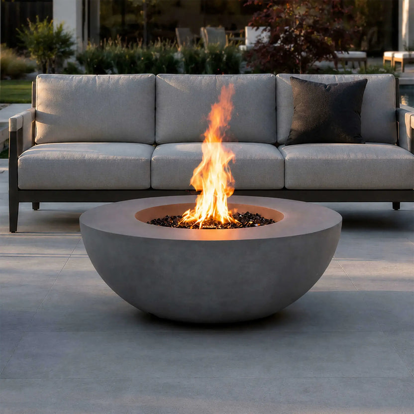 Lifestyle image of the Flame Havens Refuge Series 43-inch Round Natural Gas Gray GFRC Concrete Fire Pit burning brightly in front of a modern outdoor sofa. Reflective tempered black fire glass enhances the flames, making the fire pit a warm, elegant focal point for luxury patios, entertaining spaces, and backyard gatherings.