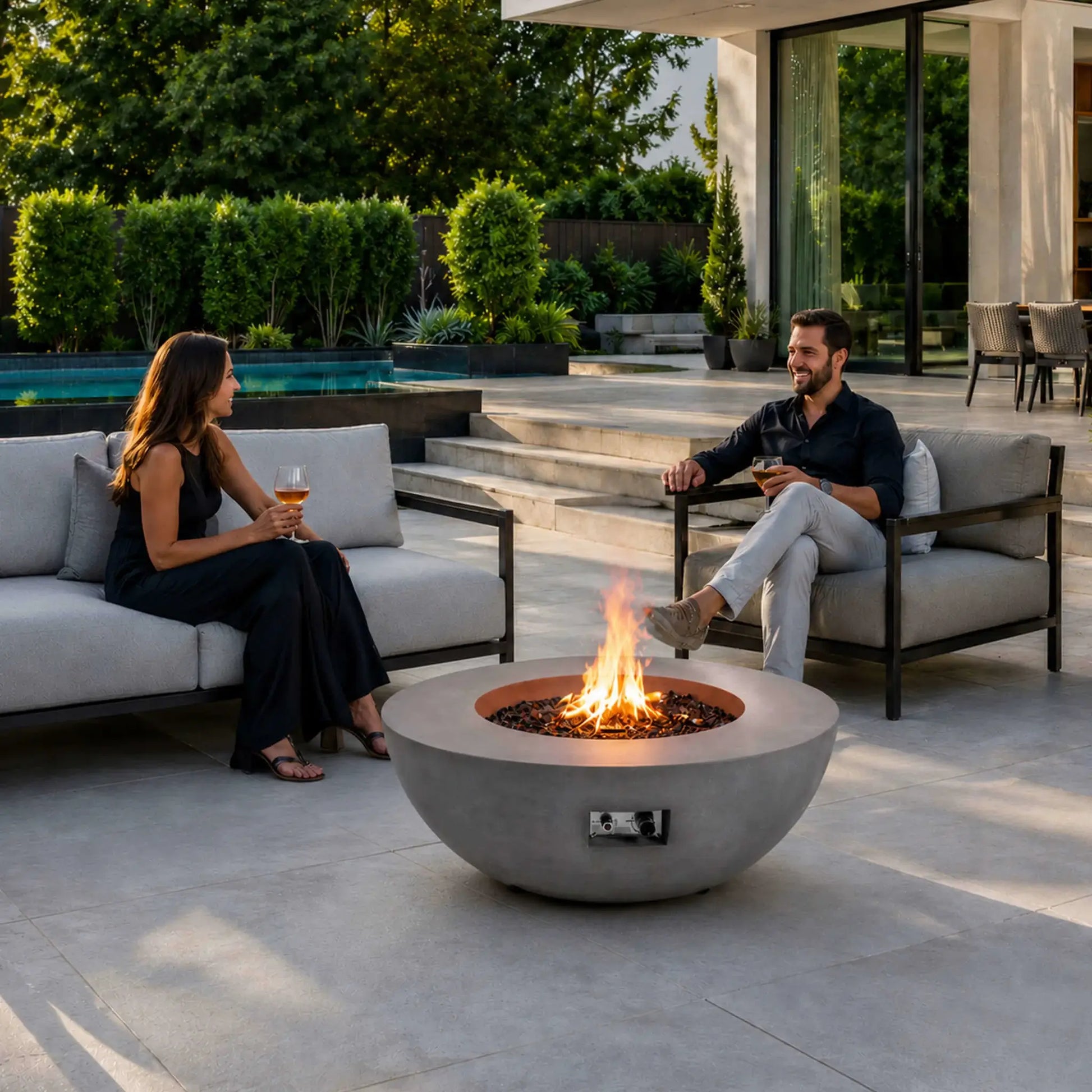 Lifestyle image of the Flame Havens Refuge Series 43-inch Round Natural Gas Gray GFRC Concrete Fire Pit with reflective tempered black fire glass and an active natural gas flame. The fire pit serves as the centerpiece of a luxury outdoor patio where two people relax with drinks, showcasing warmth, conversation, and modern outdoor living.