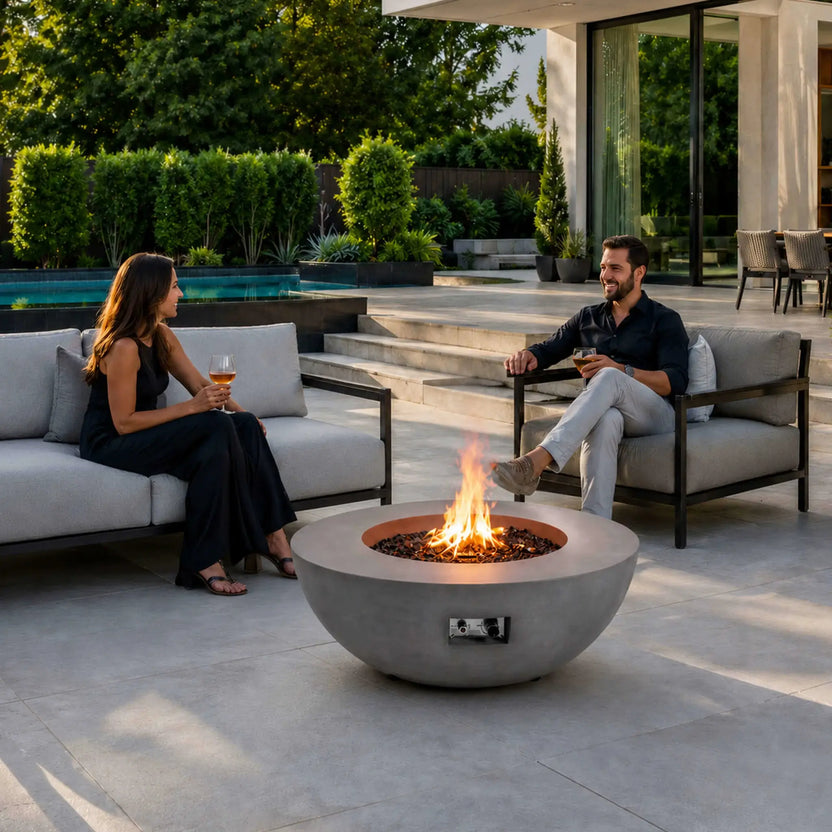 Lifestyle image of the Flame Havens Refuge Series 43-inch Round Natural Gas Gray GFRC Concrete Fire Pit with reflective tempered black fire glass and an active natural gas flame. The fire pit serves as the centerpiece of a luxury outdoor patio where two people relax with drinks, showcasing warmth, conversation, and modern outdoor living.