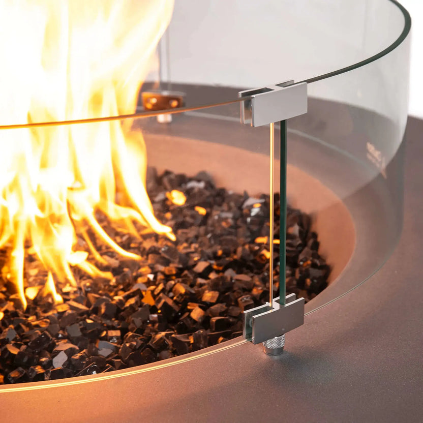 Detailed close-up of the Flame Havens Refuge Series tempered glass wind guard mounted on the 43-inch Round Natural Gas Gray GFRC Concrete Fire Pit. Stainless steel mounting brackets securely hold the curved tempered glass above the reflective tempered black fire glass while the natural gas flame burns brightly inside.