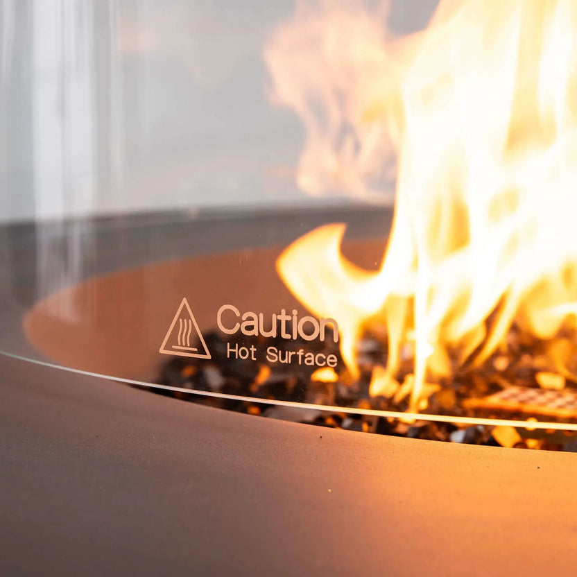 Macro view of the Flame Havens Refuge Series tempered glass wind guard displaying the etched "Caution Hot Surface" safety marking. Behind the clear glass, reflective tempered black fire glass and a vivid natural gas flame create an elegant focal point while emphasizing the premium safety features of the wind guard.