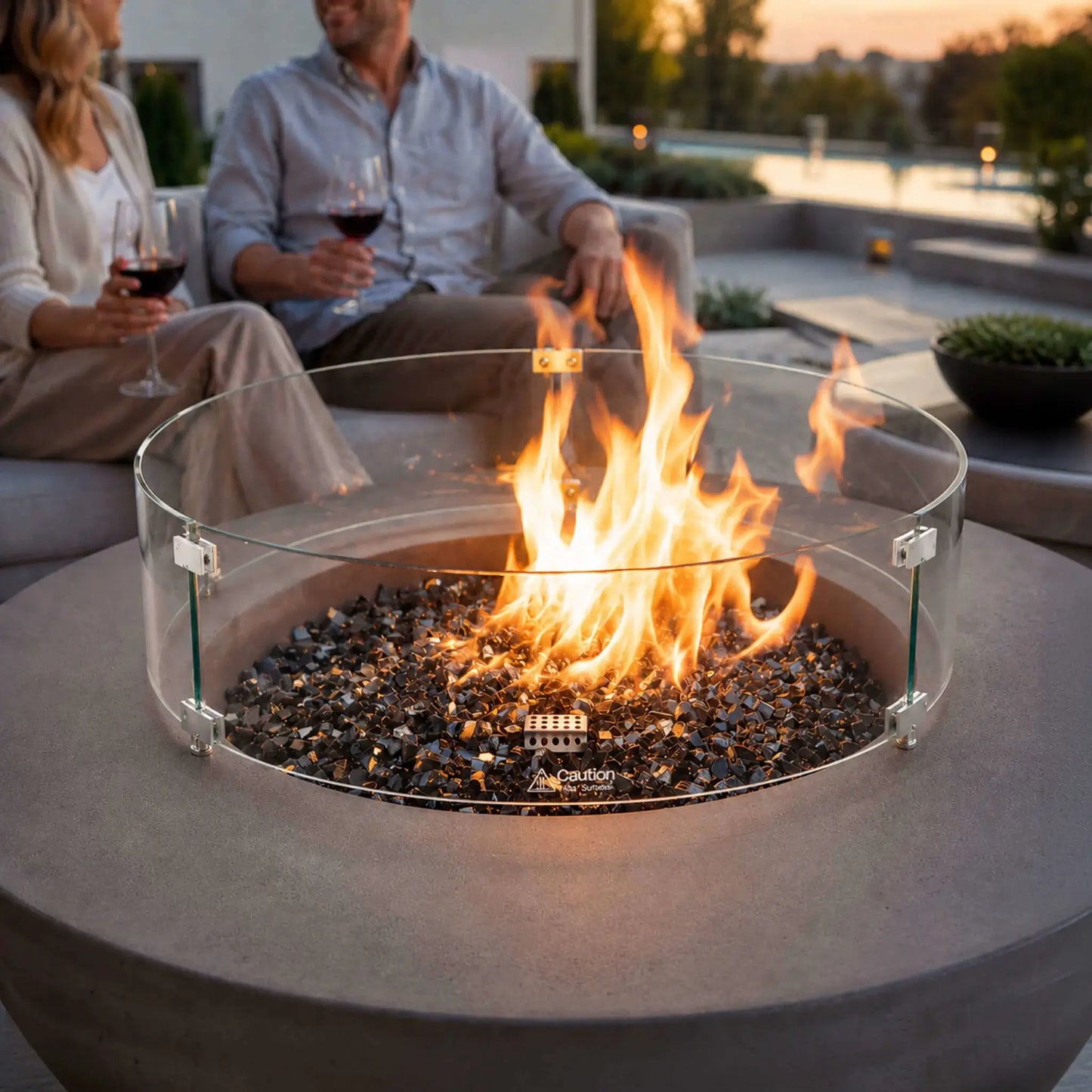 Lifestyle image of the Flame Havens Refuge Series 43-inch Round Natural Gas Gray GFRC Concrete Fire Pit equipped with a tempered glass wind guard. Reflective tempered black fire glass surrounds the lively flame as guests relax nearby with wine, demonstrating the fire pit as a stylish and inviting centerpiece for upscale outdoor entertaining.