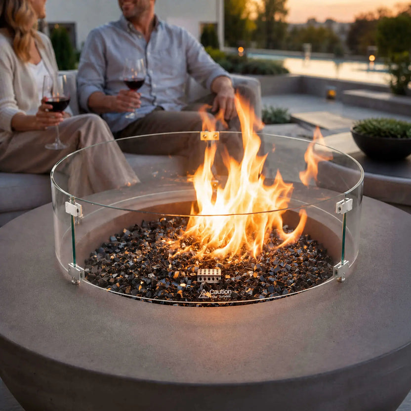 Lifestyle image of the Flame Havens Refuge Series 43-inch Round Natural Gas Gray GFRC Concrete Fire Pit equipped with a tempered glass wind guard. Reflective tempered black fire glass surrounds the lively flame as guests relax nearby with wine, demonstrating the fire pit as a stylish and inviting centerpiece for upscale outdoor entertaining.