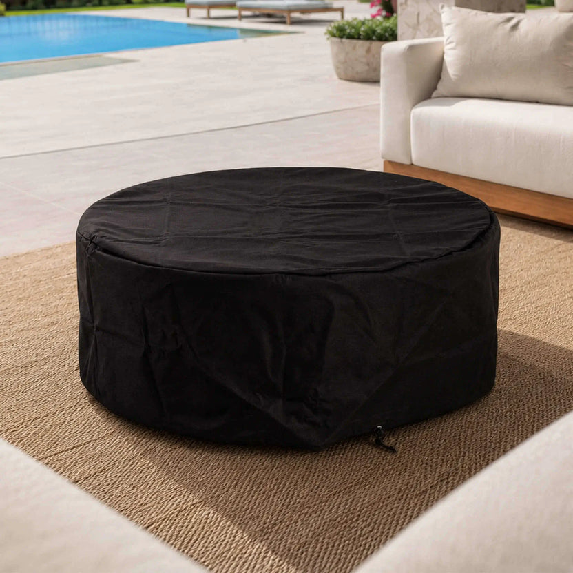 Lifestyle image of the Flame Havens Refuge Series 43-inch Round Natural Gas White GFRC Concrete Fire Pit covered with its black canvas fabric cover on a luxury outdoor patio beside a swimming pool. The fitted cover keeps the fire pit clean and protected while blending seamlessly into the modern outdoor living space.