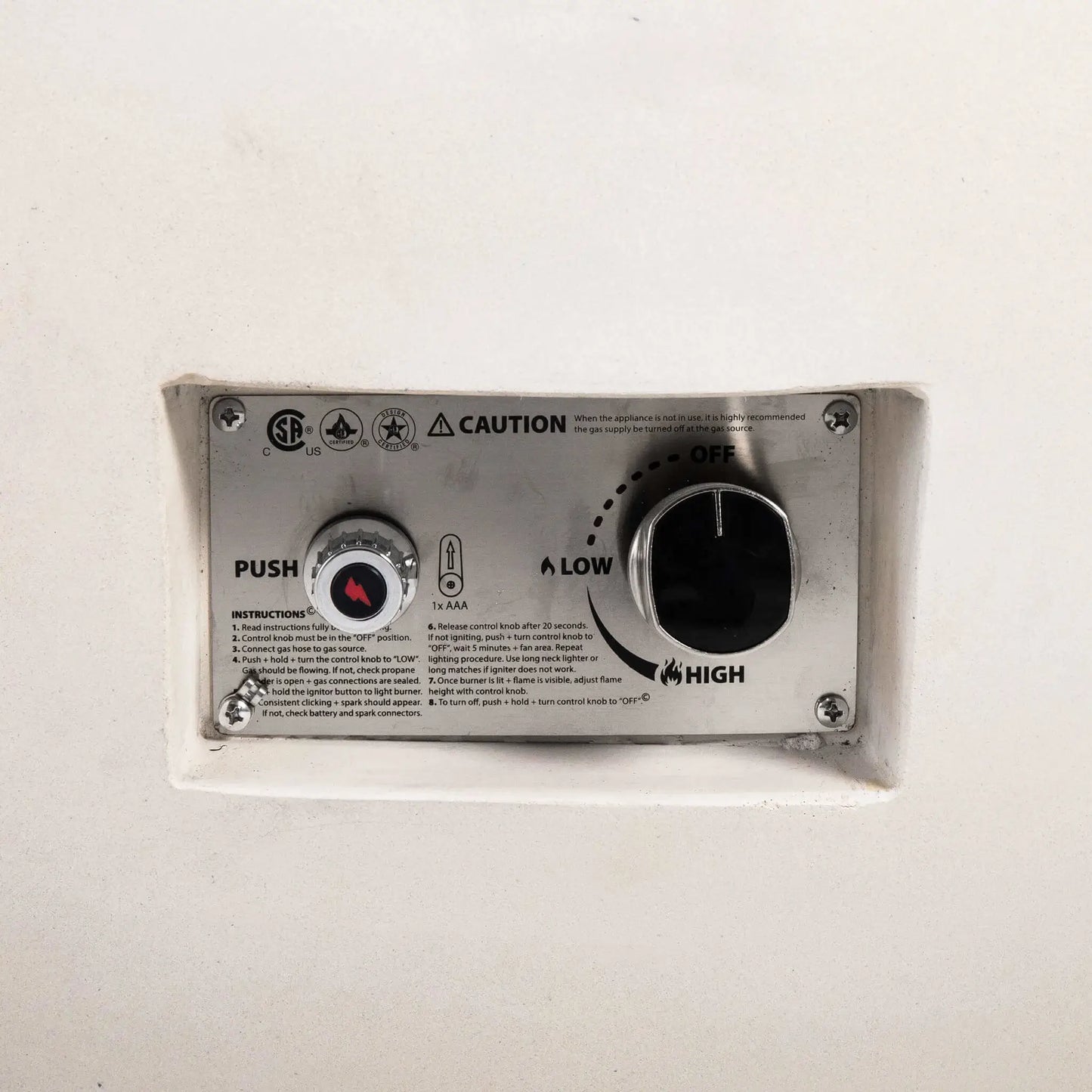 Close-up of the recessed control panel on the Flame Havens Refuge Series 43-inch Round Natural Gas White GFRC Concrete Fire Pit. The image clearly shows the push-button electronic ignition, flame control knob, ignition instructions, and CSA-certified safety labeling integrated into the fire pit's clean exterior.