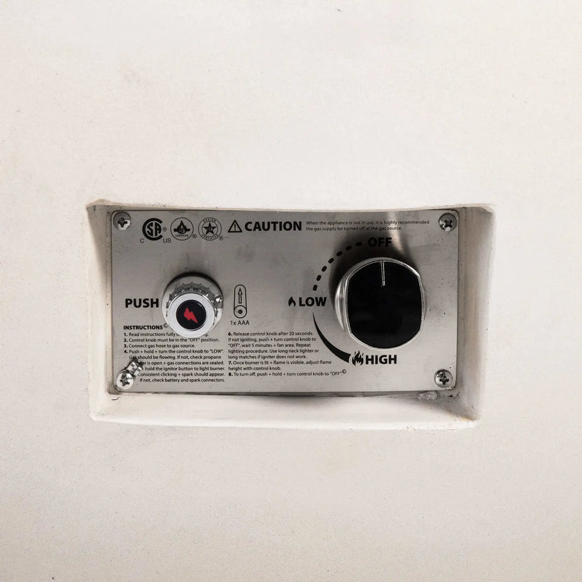 Close-up of the recessed control panel on the Flame Havens Refuge Series 43-inch Round Natural Gas White GFRC Concrete Fire Pit. The image clearly shows the push-button electronic ignition, flame control knob, ignition instructions, and CSA-certified safety labeling integrated into the fire pit's clean exterior.