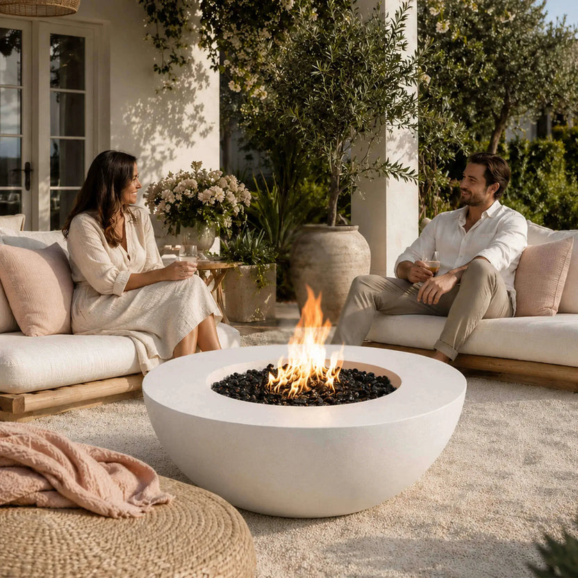 Lifestyle scene featuring the Flame Havens Refuge Series 43-inch Round Natural Gas White GFRC Concrete Fire Pit with reflective tempered black fire glass burning brightly between two guests relaxing on a covered patio. The inviting fire creates a warm centerpiece for conversation in an elegant outdoor living space surrounded by greenery.