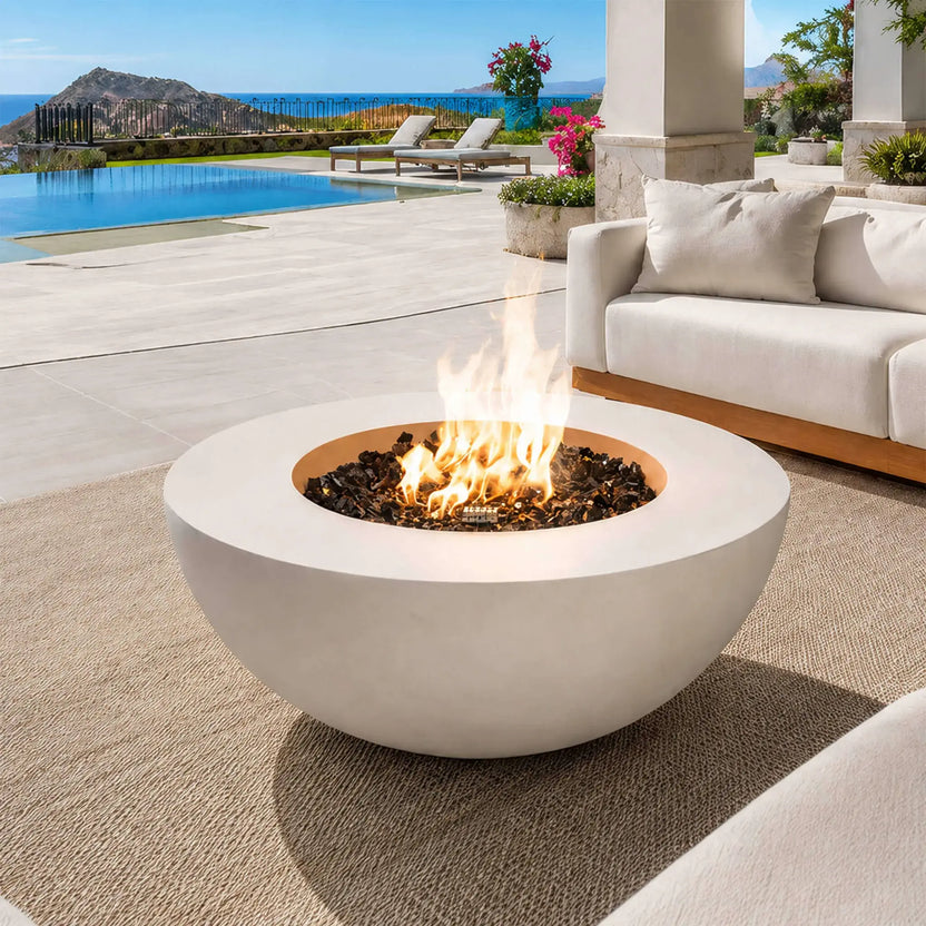 The Flame Havens Refuge Series 43-inch Round Natural Gas White GFRC Concrete Fire Pit creates a stunning focal point on a luxury outdoor patio overlooking an infinity pool and ocean view. Reflective tempered black fire glass and tall flames provide warmth while complementing modern outdoor seating and upscale resort-style architecture.