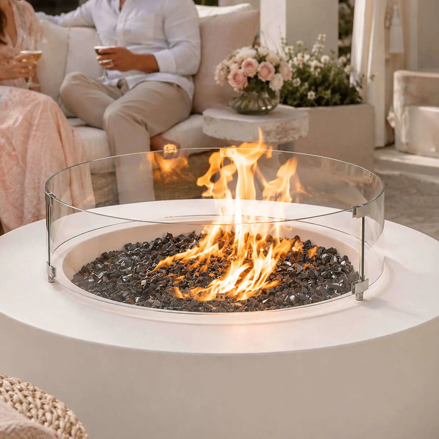 Lifestyle image of the Flame Havens Refuge Series 43-inch Round Natural Gas White GFRC Concrete Fire Pit featuring a tempered glass wind guard, reflective tempered black fire glass, and lively natural gas flames. Friends relax around the fire on a luxurious outdoor patio, highlighting the fire pit as a stylish centerpiece for entertaining and year-round outdoor living.