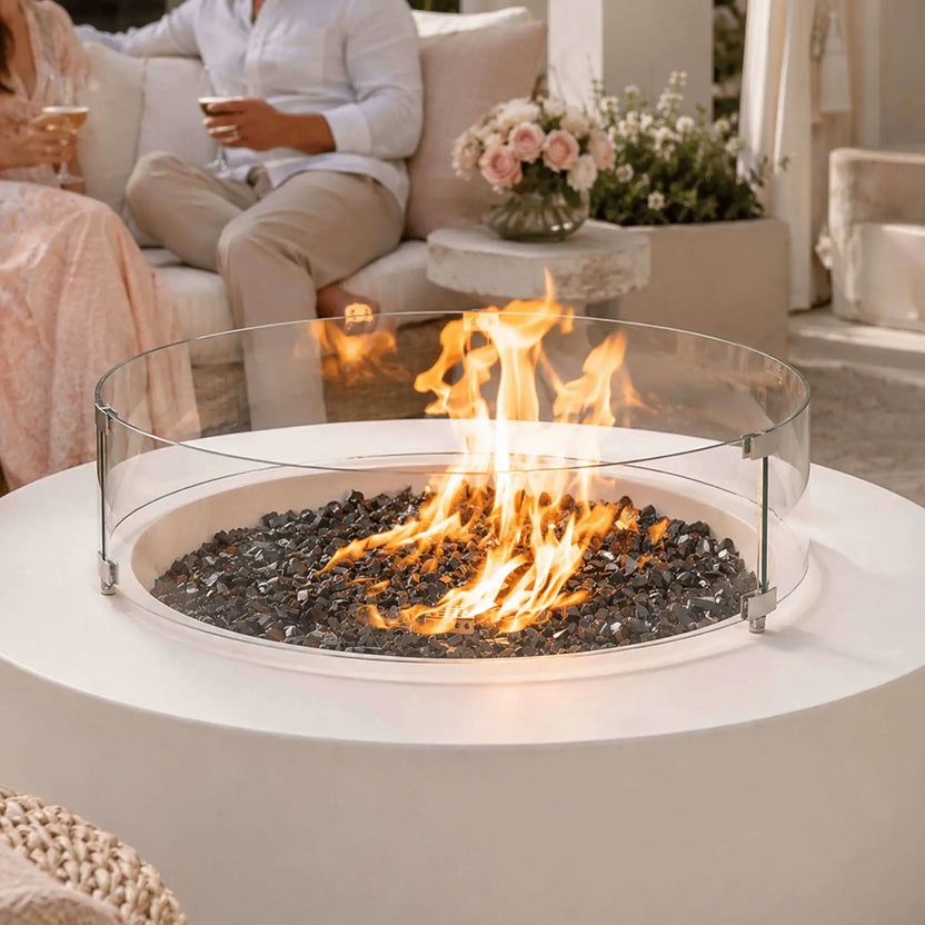 Lifestyle image of the Flame Havens Refuge Series 43-inch Round Natural Gas White GFRC Concrete Fire Pit featuring a tempered glass wind guard, reflective tempered black fire glass, and lively natural gas flames. Friends relax around the fire on a luxurious outdoor patio, highlighting the fire pit as a stylish centerpiece for entertaining and year-round outdoor living.