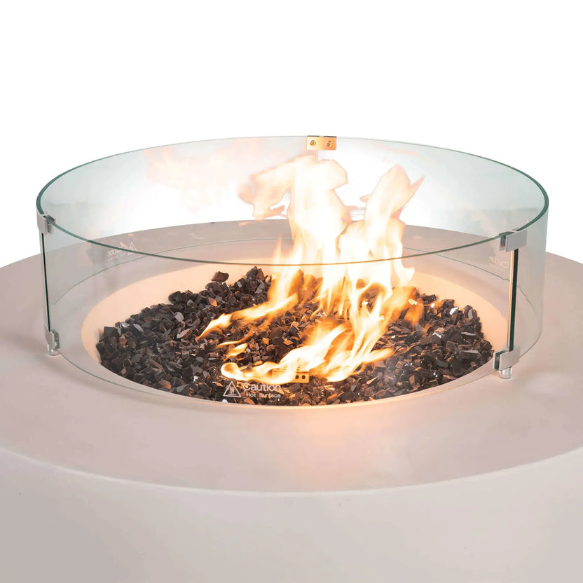 The Flame Havens Refuge Series 43-inch Round Natural Gas White GFRC Concrete Fire Pit features a clear tempered glass wind guard surrounding reflective tempered black fire glass and vibrant natural gas flames. The transparent wind guard provides added protection while preserving unobstructed views of the fire from every angle.