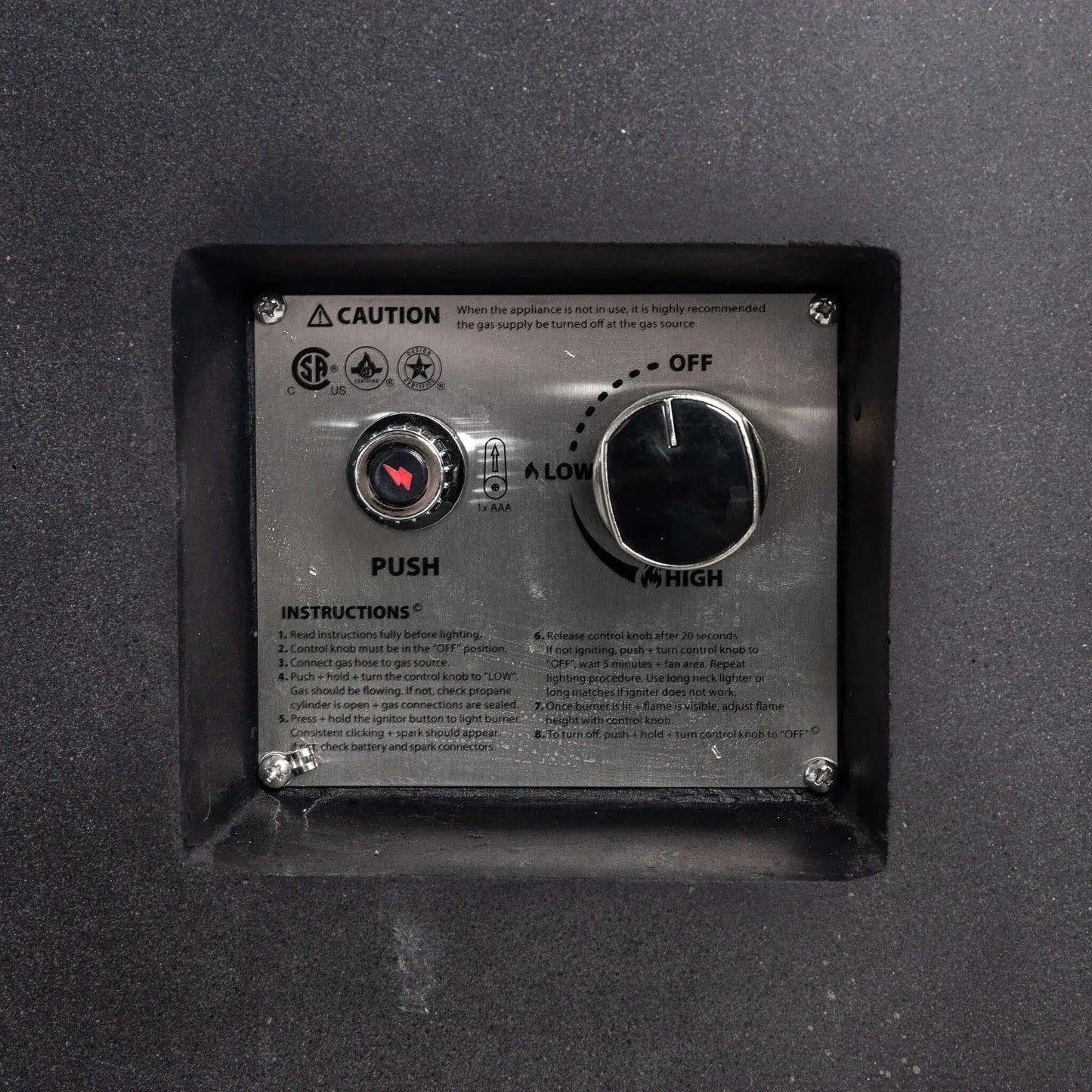Close-up of the Flame Havens Refuge Series 43-inch Round Propane Gas GFRC Concrete Fire Pit recessed CSA-certified ignition control panel. The image showcases the electronic push-button igniter, flame adjustment control knob, operating instructions, and stainless steel control plate neatly integrated into the black GFRC concrete fire pit for convenient operation.