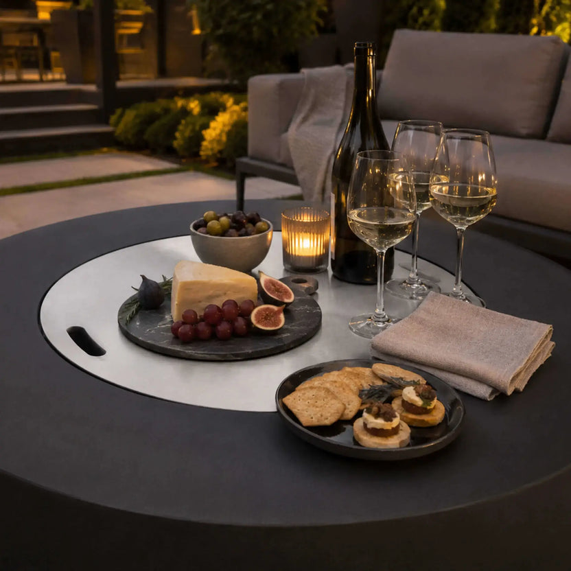 Lifestyle image of the Flame Havens Refuge Series 43-inch Round Propane Gas GFRC Concrete Fire Pit in Black with the optional stainless steel burner cover installed. Styled as an outdoor entertaining table with wine, cheese, fruit, crackers, and candlelight, the covered fire pit demonstrates its versatility as both a luxury fire feature and elegant patio gathering space.