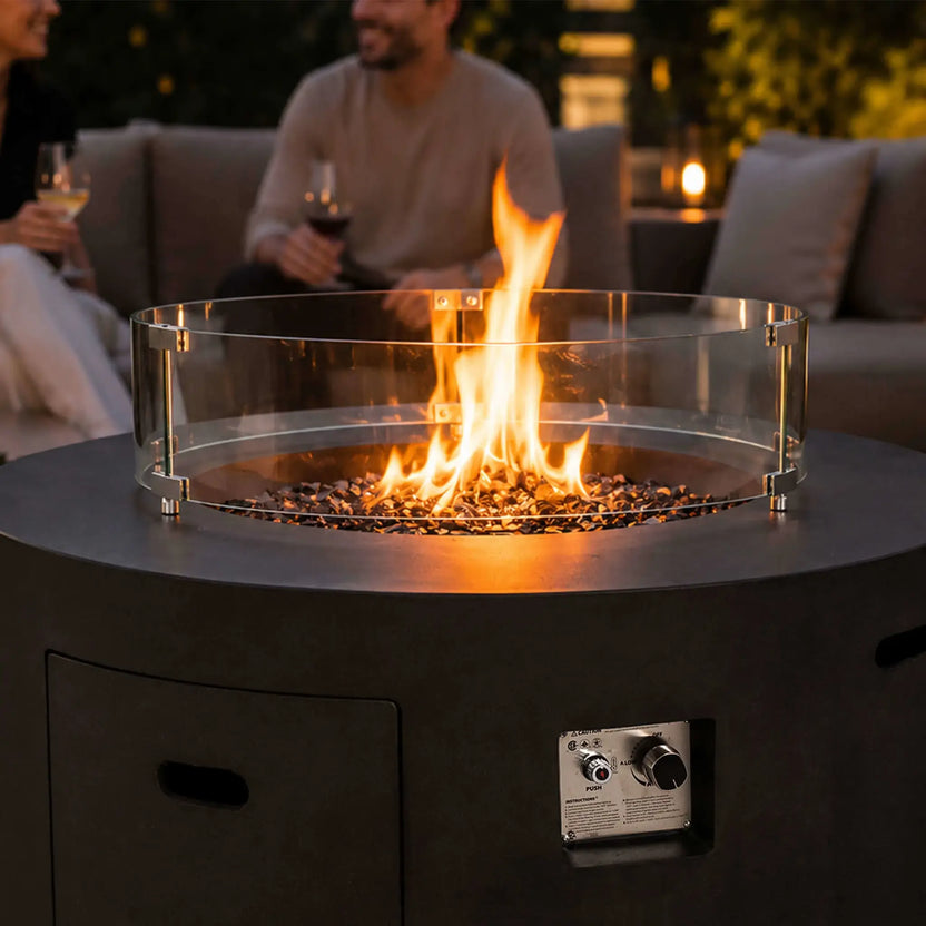 Lifestyle image of the Flame Havens Refuge Series 43-inch Round Propane Gas GFRC Concrete Fire Pit in Black featuring the optional tempered glass wind guard and a lively propane flame above black reflective fire glass. Set within a sophisticated outdoor seating area, the fire pit creates a warm gathering space while showcasing its premium GFRC construction and modern design.