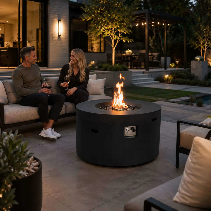 Lifestyle image of the Flame Havens Refuge Series 43-inch Round Propane Gas GFRC Concrete Fire Pit in Black with a lively flame burning above black reflective fire glass. Positioned in a modern luxury backyard, the fire pit creates a welcoming gathering space where two people relax with wine, showcasing the hidden propane design and contemporary outdoor entertaining appeal.