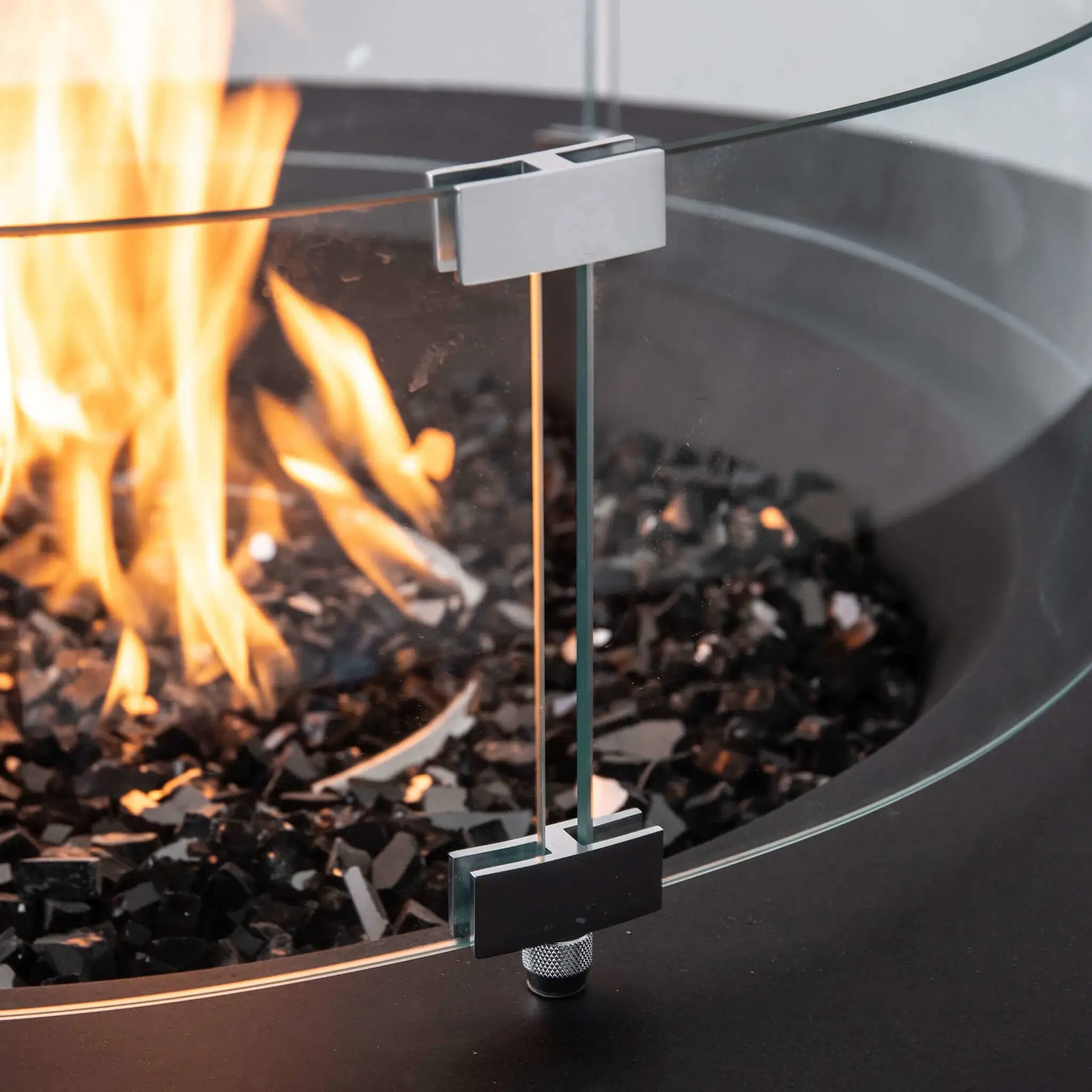 Close-up of the optional tempered glass wind guard installed on the Flame Havens Refuge Series 43-inch Round Propane Gas GFRC Concrete Fire Pit in Black. The image highlights the premium stainless steel mounting brackets, crystal-clear tempered glass panels, black reflective fire glass, and active propane flame, demonstrating the wind guard's secure construction and elegant finish.