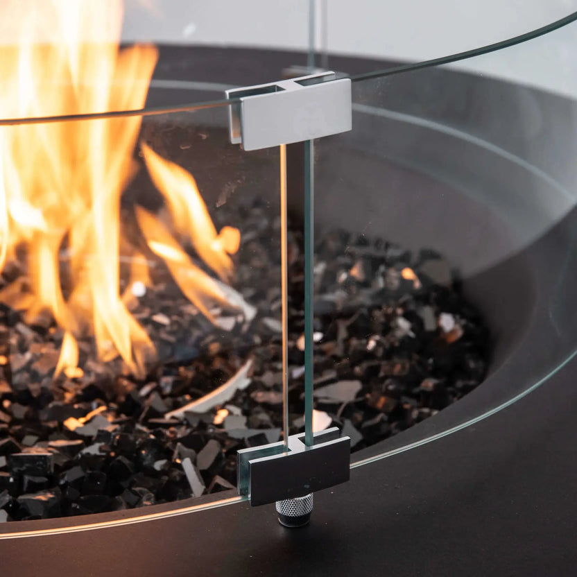 Close-up of the optional tempered glass wind guard installed on the Flame Havens Refuge Series 43-inch Round Propane Gas GFRC Concrete Fire Pit in Black. The image highlights the premium stainless steel mounting brackets, crystal-clear tempered glass panels, black reflective fire glass, and active propane flame, demonstrating the wind guard's secure construction and elegant finish.
