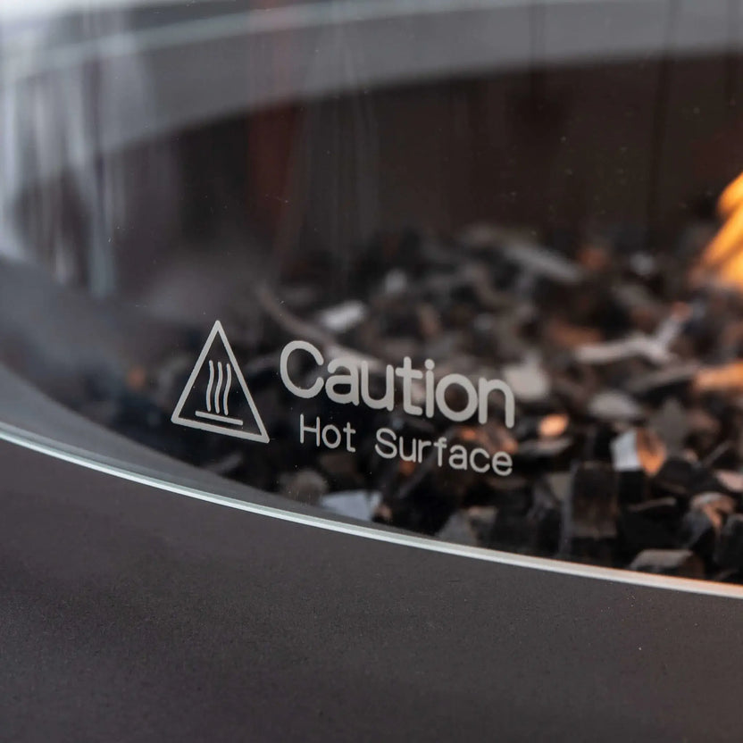 Detailed close-up of the tempered glass wind guard on the Flame Havens Refuge Series 43-inch Round Propane Gas GFRC Concrete Fire Pit in Black. The etched "Caution Hot Surface" safety marking is clearly visible on the glass, with black reflective fire glass and a warm propane flame softly blurred in the background, showcasing the premium safety features and refined craftsmanship.