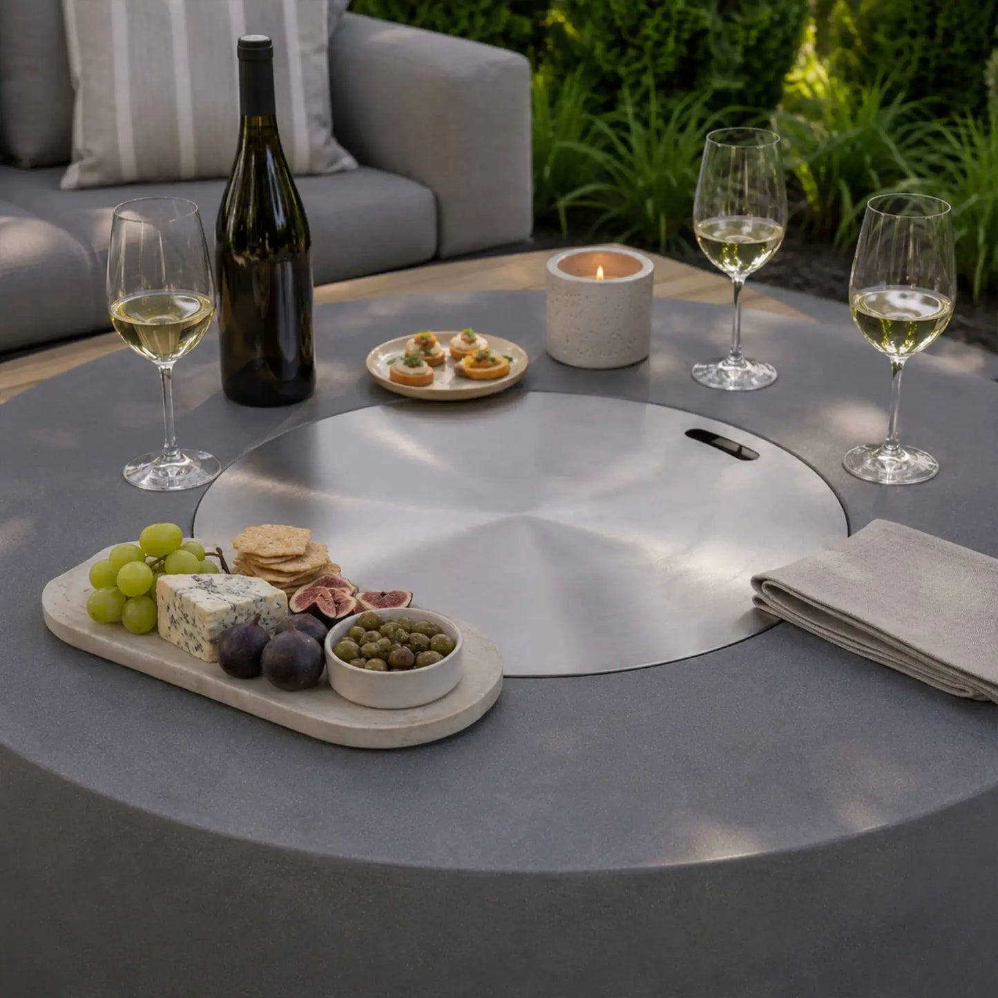 Lifestyle image of the Flame Havens Refuge Series 43-inch Round Propane Gas GFRC Concrete Fire Pit in Gray with the stainless steel burner cover installed, transforming the fire pit into a functional outdoor table. The surface is styled with wine glasses, a bottle of wine, charcuterie board, appetizers, candles, and folded napkins, showcasing elegant outdoor entertaining.