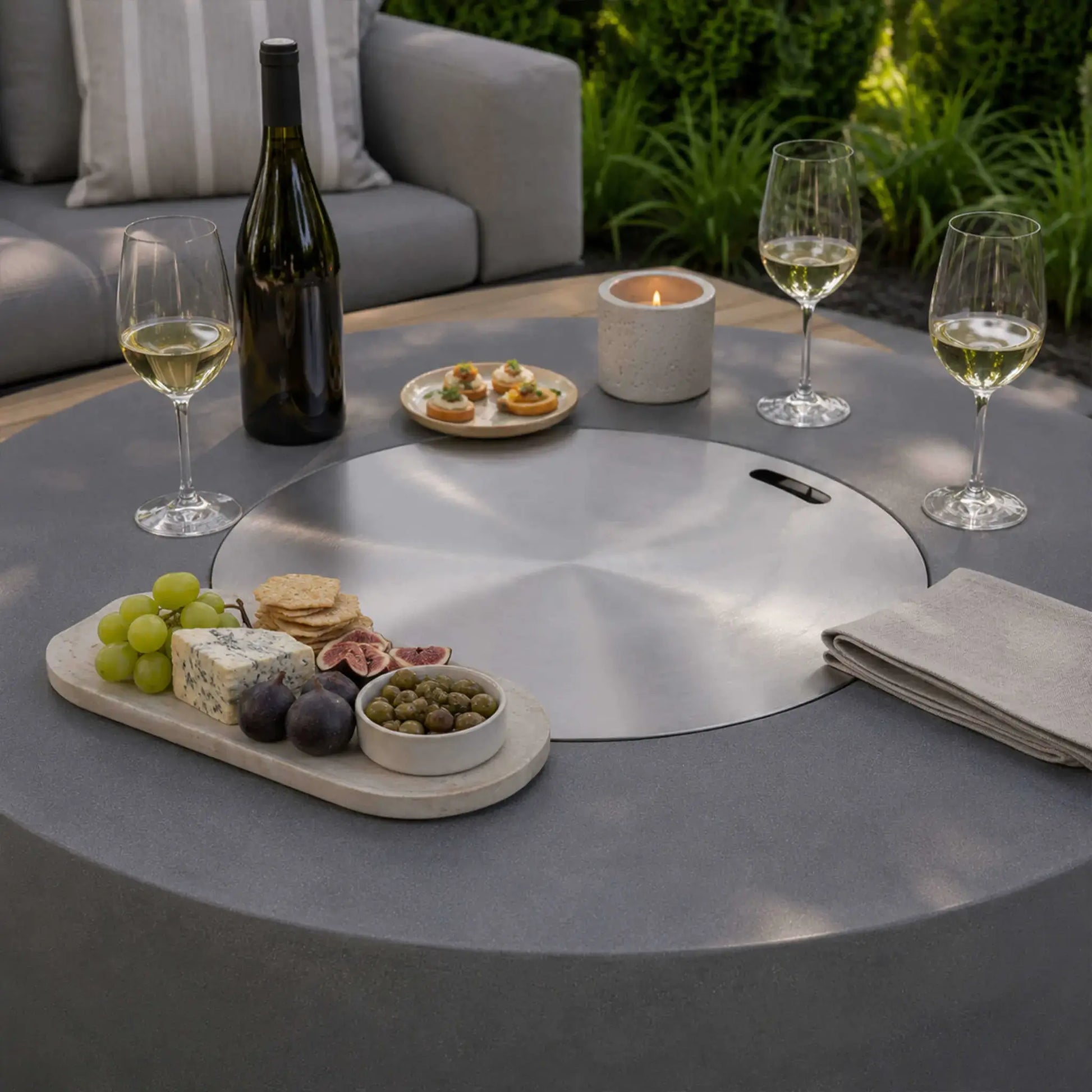 Lifestyle image of the Flame Havens Refuge Series 43-inch Round Propane Gas GFRC Concrete Fire Pit in Gray with the stainless steel burner cover installed, transforming the fire pit into a functional outdoor table. The surface is styled with wine glasses, a bottle of wine, charcuterie board, appetizers, candles, and folded napkins, showcasing elegant outdoor entertaining.