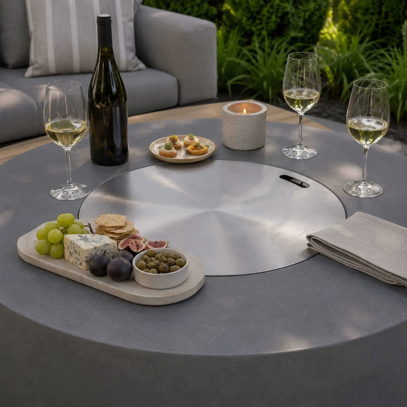 Lifestyle image of the Flame Havens Refuge Series 43-inch Round Propane Gas GFRC Concrete Fire Pit in Gray with the stainless steel burner cover installed, transforming the fire pit into a functional outdoor table. The surface is styled with wine glasses, a bottle of wine, charcuterie board, appetizers, candles, and folded napkins, showcasing elegant outdoor entertaining.