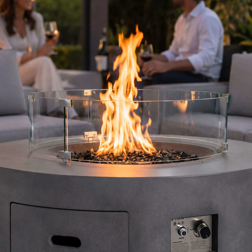 Lifestyle image of the Flame Havens Refuge Series 43-inch Round Propane Gas GFRC Concrete Fire Pit in Gray featuring premium black reflective fire glass and the optional tempered glass wind guard. A vibrant flame creates a warm focal point while guests relax in a luxury outdoor seating area, demonstrating the fire pit's elegant design for upscale backyard entertaining.