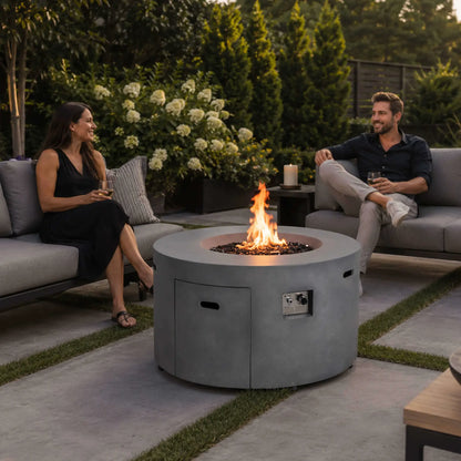 Lifestyle image of the Flame Havens Refuge Series 43-inch Round Propane Gas GFRC Concrete Fire Pit in Gray with tempered black reflective fire glass burning brightly. The fire pit is centered between luxury outdoor sofas where two guests enjoy drinks and conversation, demonstrating how the propane fire pit creates a warm and inviting atmosphere for premium backyard entertaining.