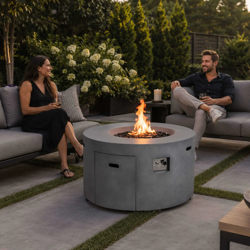 Lifestyle image of the Flame Havens Refuge Series 43-inch Round Propane Gas GFRC Concrete Fire Pit in Gray with tempered black reflective fire glass burning brightly. The fire pit is centered between luxury outdoor sofas where two guests enjoy drinks and conversation, demonstrating how the propane fire pit creates a warm and inviting atmosphere for premium backyard entertaining.